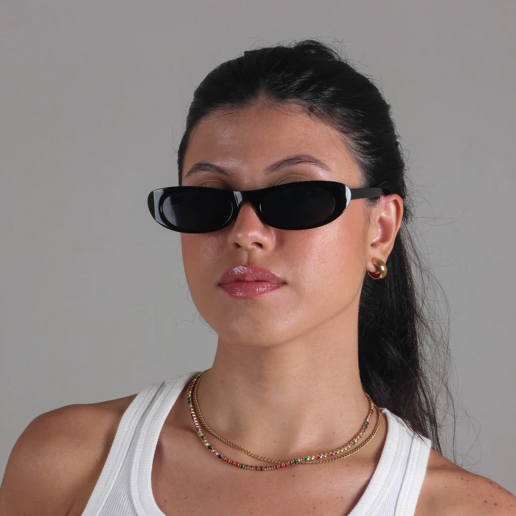 Woman wearing Nova oval slim sunglasses in Black frame with Black acrylic lenses, polycarbonate frame, 100% UV protection, facing camera with head straight, hands not visible, white wall background.
