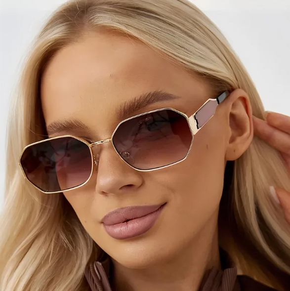Model wearing Luna women’s retro geometric shades in Gold frame with Brown Gradient lenses, in front of white wall, head slightly tilted left, looking at the camera, right hand placed behind right ear