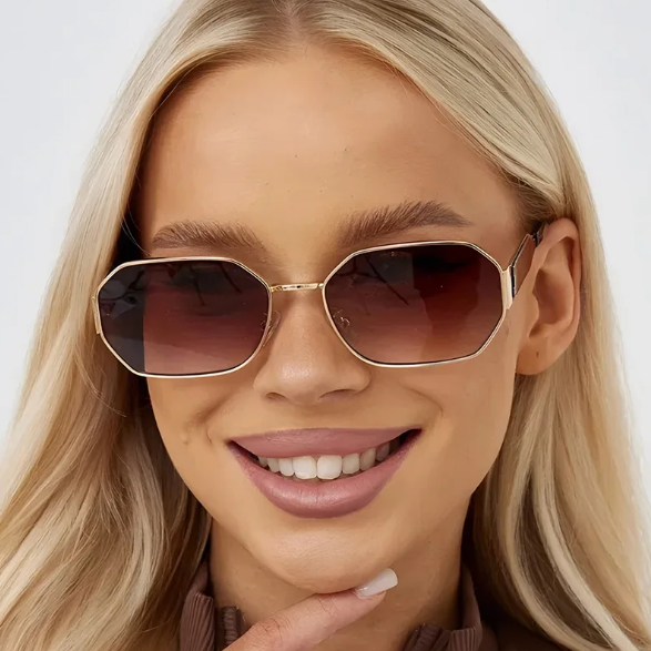 Model wearing Luna women’s retro geometric shades in Gold frame with Brown Gradient lenses, in front of white wall, facing forward and looking at the camera with a slight smile, left hand touching chin