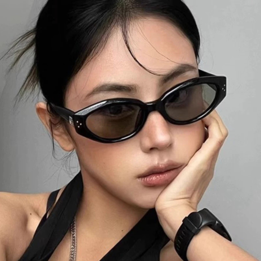 Serena women’s oval slim sunglasses in Black frame with Black lenses, worn by model against light gray wall — model facing forward, looking at camera with right hand resting on chin — premium acetate frame, Trivex lenses, 100% UV protection