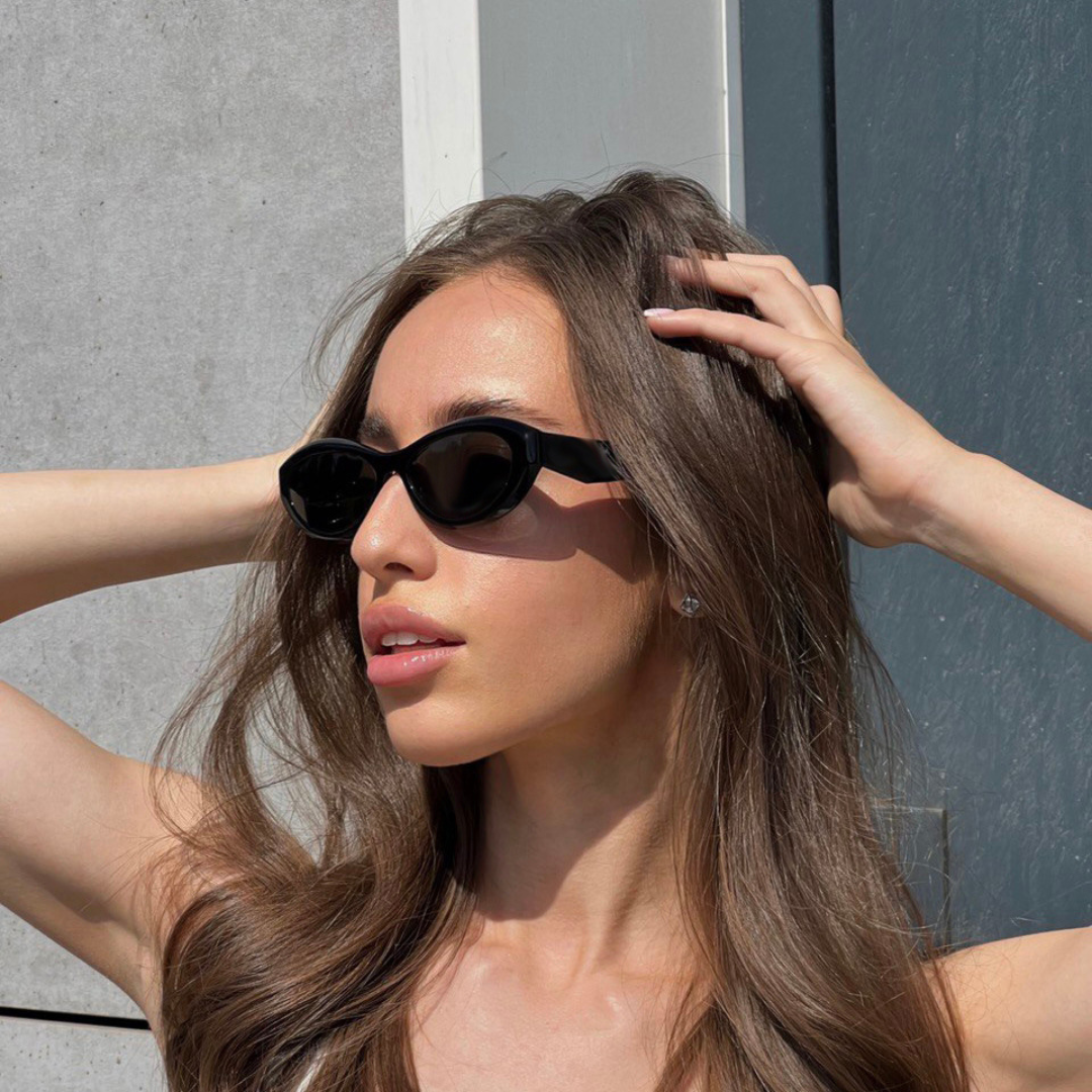 Woman wearing Liora cat eye  shades background in Black acetate frame with black TAC lenses, 100% UV protection, modern concrete wall and blue-gray metal door background, looking to the left with both hands intertwined in hair