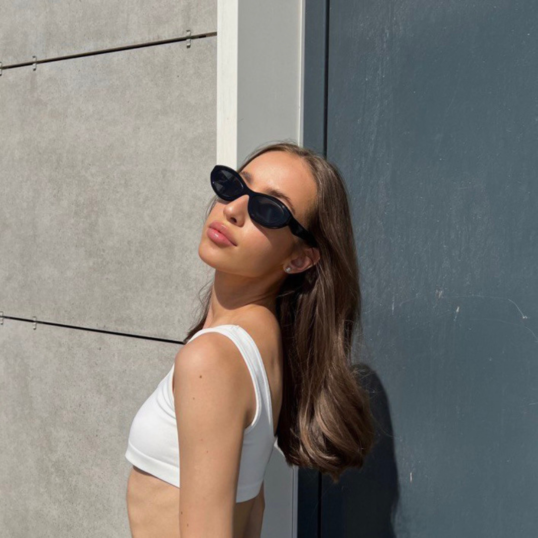Woman wearing Liora cat eye sunglasses in Black acetate frame with black TAC lenses, 100% UV protection, modern concrete wall and blue-gray metal door background, body turned to the left while looking forward, hands relaxed