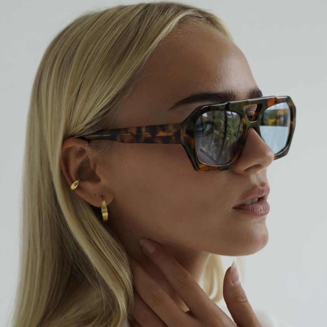 Woman wearing Ravena aviator shades in Tortoise frame with soft blue lenses, white wall background, looking almost fully to the right with hand touching neck