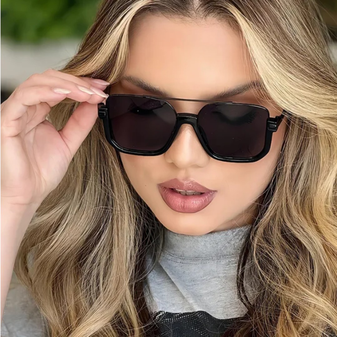 Alt text: Model wearing Opéra women's aviator-inspired square sunglasses, black lightweight metal frame with black UV400 resin lenses, facing forward with chin slightly lowered, looking slightly down, left hand touching the frame, outdoor background.