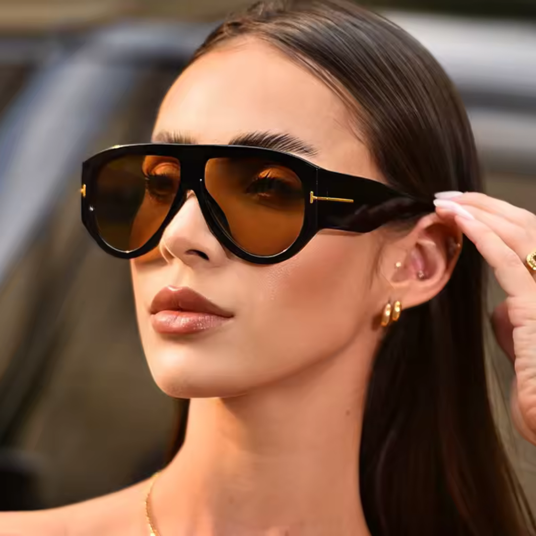 Woman wearing Cannes oversized shield sunglasses in black acetate frame with amber TAC lenses, 100% UV protection, outdoor setting, face turned slightly left looking slightly to the left, right hand touching the sunglasses frame