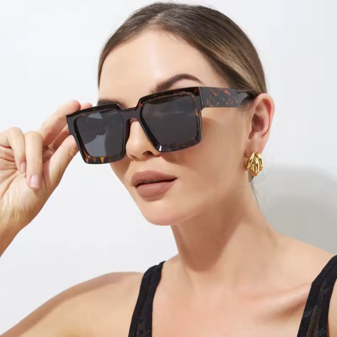 Valencia women’s square sunglasses in Black frame with Black lenses, worn by model against white wall — model facing slightly left, looking left, left hand touching the left side of the sunglasses frame — acetate frame, polycarbonate lenses, 100% UV protection