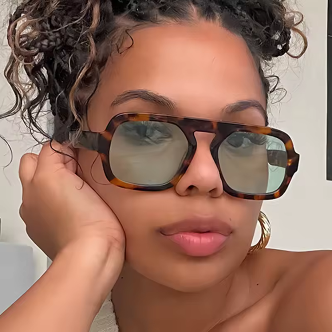 Model wearing Zara women’s shades in Tortoise frame with Dark Green lenses, against light beige wall, facing forward and looking at the camera with left hand touching left side of face