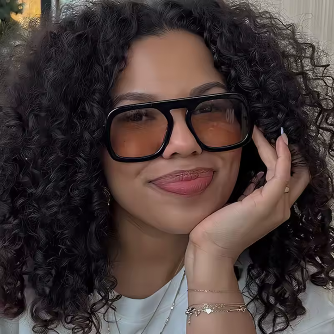 Model wearing Zara women’s sunglasses in Black frame with Amber lenses, outdoors, facing forward and looking at the camera with a slight smile, right hand resting on chin