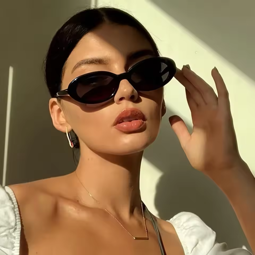 Woman wearing Amélie oval sunglasses in Black polycarbonate frame with black acrylic lenses, 100% UV protection, light beige wall background, looking at the camera with right hand touching right side of sunglasses frame
