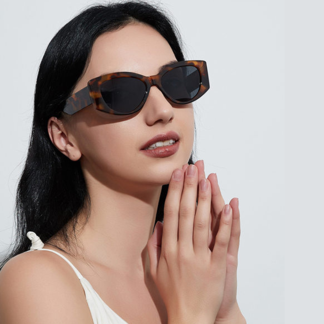 Female model wearing Aravie women’s cat-eye sunglasses in Tortoise frame with Black lenses, against a white wall, looking slightly to the right with hands together and a subtle smile