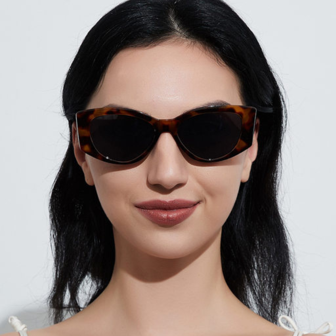 Female model wearing Aravie women’s cat-eye sunglasses in Tortoise frame with Black lenses, looking directly at the camera against white wall, hands not visible