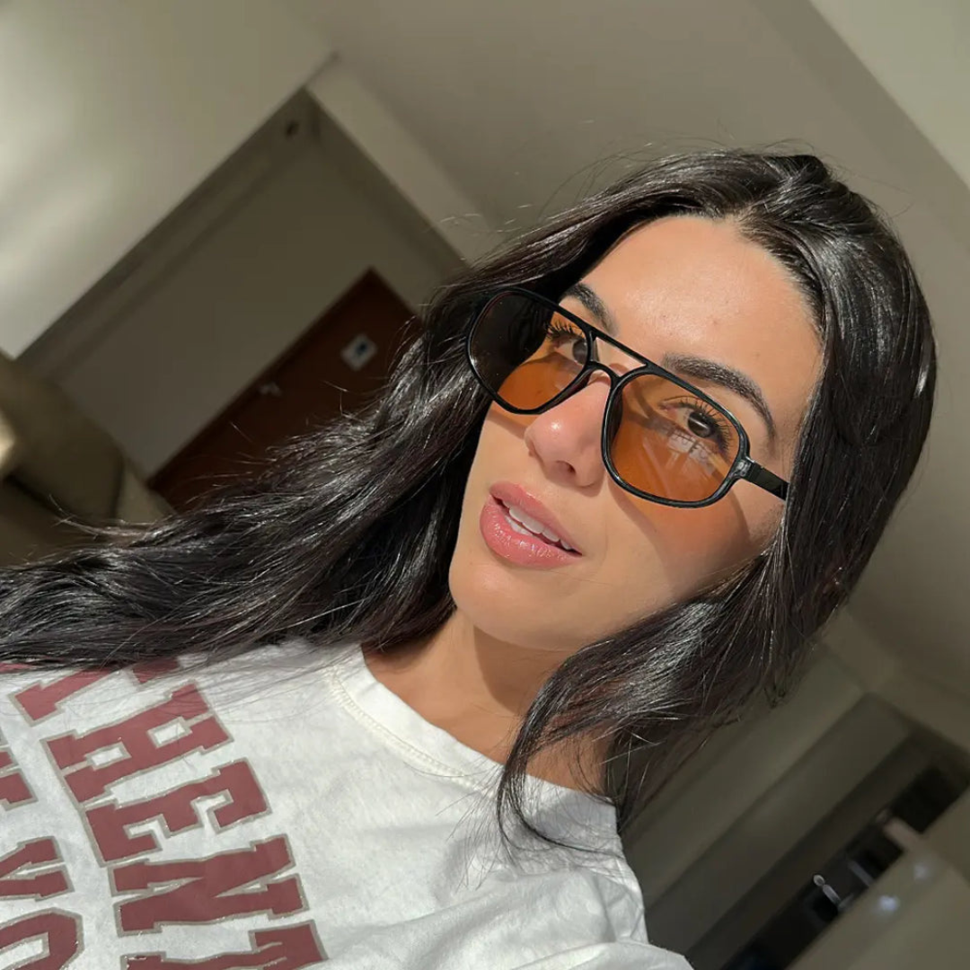 Woman wearing Brigitte aviator sunglasses in black polycarbonate frame with amber acrylic lenses, 100% UV protection, outdoor setting, face facing forward looking at the camera with a slight smile