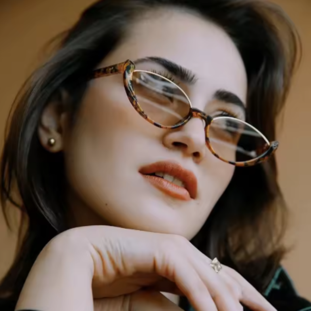Model wearing Venice women's cat-eye sunglasses, tortoise premium acetate frame with brown gradient TAC lenses offering UV400 protection, face slightly turned to the right looking upwards, right hand touching chin, beige wall background.