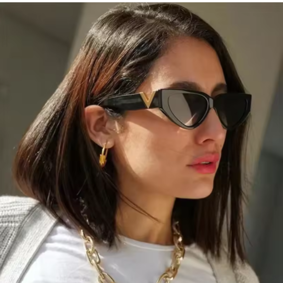 Riviera women’s cat-eye shades in Black frame with Black lenses, worn by model against concrete wall — model facing slightly right, looking slightly right, hands not visible — polycarbonate frame, acrylic lenses, 100% UV protection