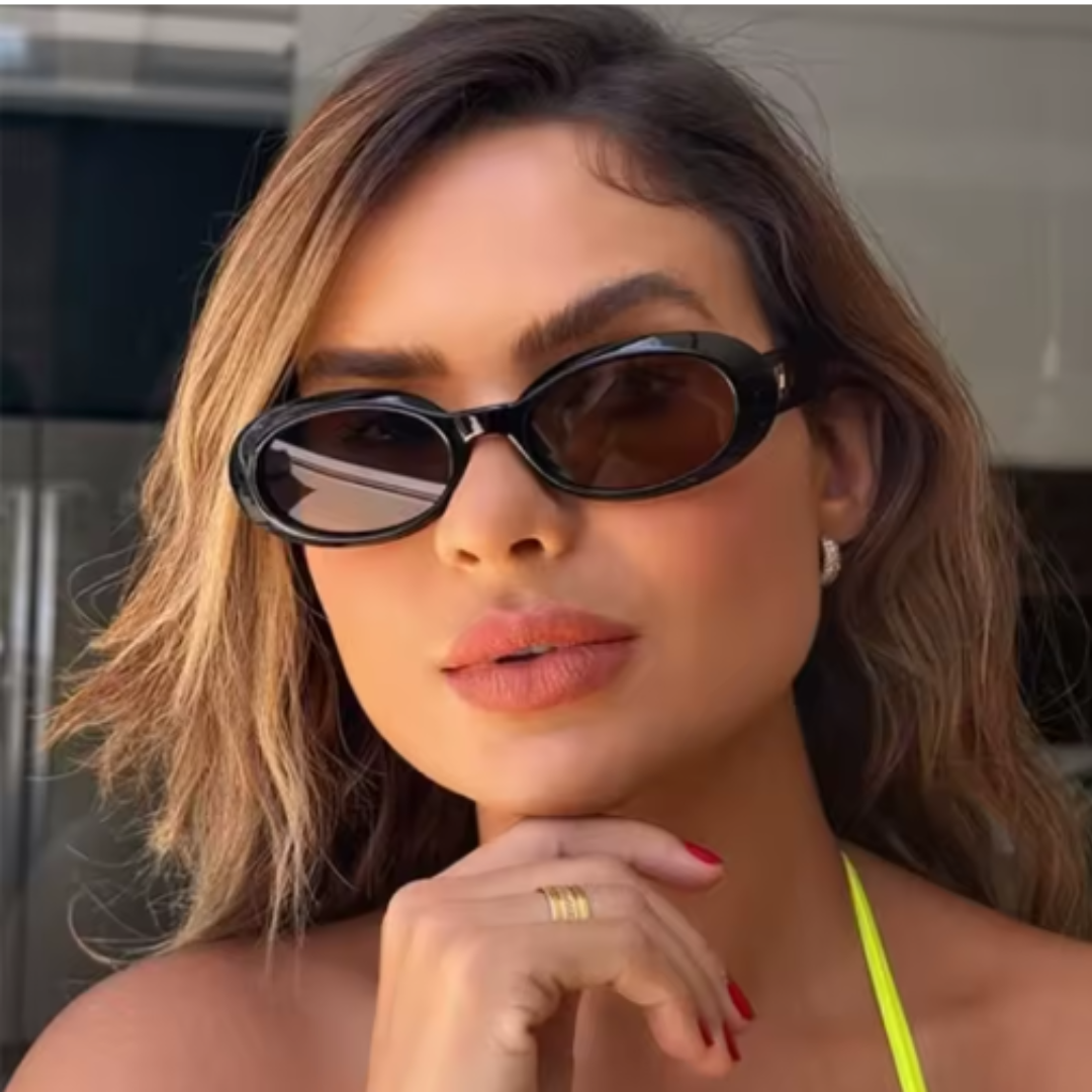Female model wearing Vienna oval sunglasses in Black frame with Black lenses, outdoor background, looking forward at the camera with left hand touching chin and a slight smile — acetate frame, polycarbonate lenses, 100% UV protection