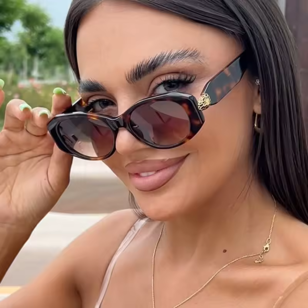 Female model wearing Selene women’s wide oval shades in Black frame with Black lenses, outdoors, head slightly turned to the left but looking over the top of the sunglasses directly at the camera and smiling, left hand touching the sunglasses frame