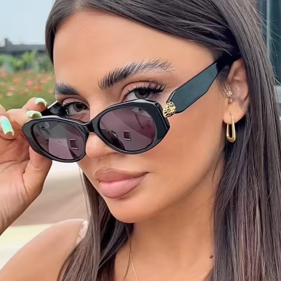 Female model wearing Selene women’s wide oval sunglasses in Black frame with Black lenses, outdoors, head slightly turned to the left but looking over the top of the sunglasses directly at the camera, left hand touching the sunglasses frame
