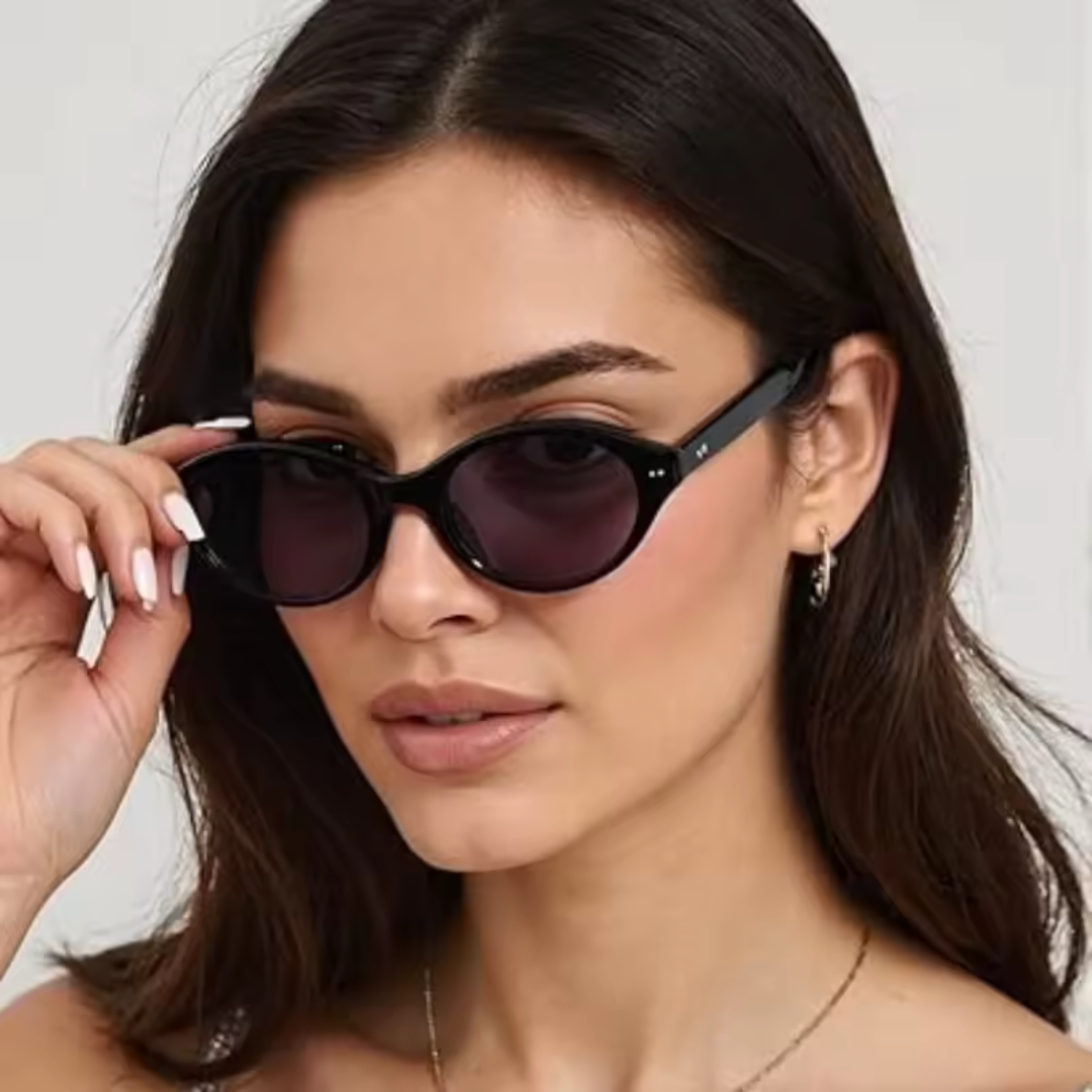 Female model wearing Sovereign women’s oval slim sunglasses in Black frame with Black lenses, standing against light beige concrete wall, head slightly turned to the left but looking directly at the camera, left hand touching the sunglasses frame