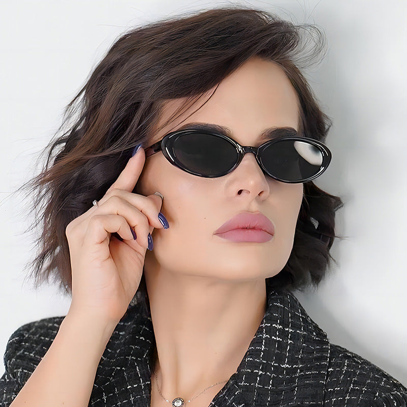 Model wearing Évara oval women's shades in Black polycarbonate frame with black polycarbonate lenses, 100% UV protection, facing slightly right with left hand touching the left temple, white wall background