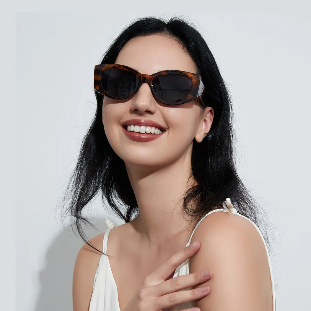 Female model wearing Aravie women’s cat-eye  shades in Tortoise frame with Black lenses, looking directly at the camera with left hand touching shoulder and smiling, right hand not visible, white wall background