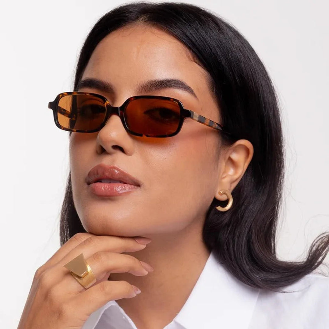 Model wearing Zayla rectangular women's sunglasses in Leopard polycarbonate frame with amber polycarbonate lenses, 100% UV protection, face turned slightly left looking at camera with hand gently touching chin, white wall background