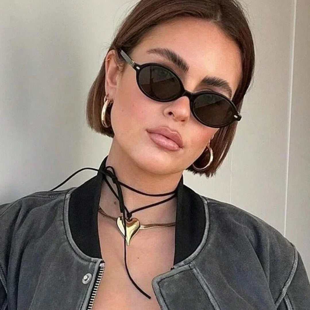 Female model wearing Lunéra women’s oval shades in Black frame with Black lenses, looking directly at the camera with head slightly tilted to the right, beige wall background, hands not visible