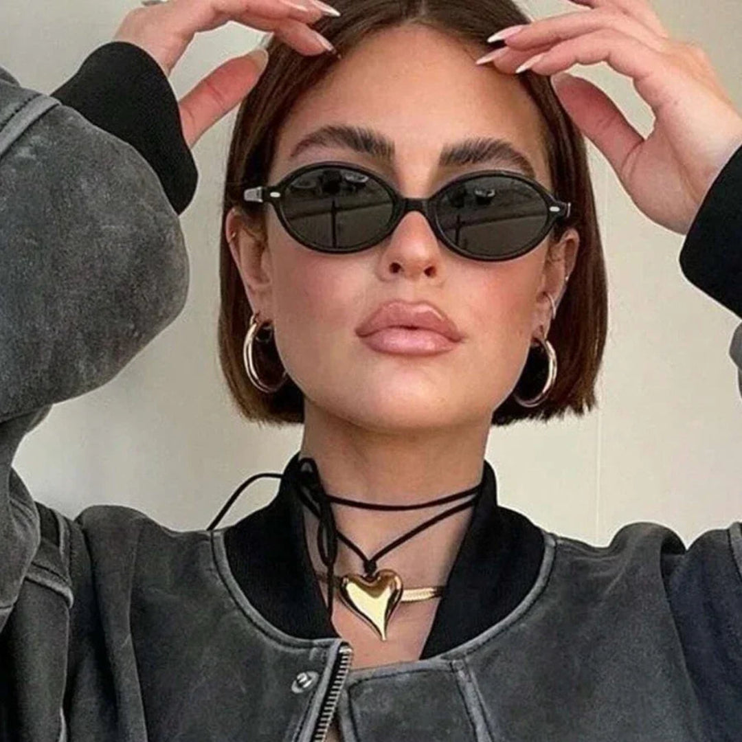 Female model wearing Lunéra women’s oval sunglasses in Black frame with Black lenses, looking directly at the camera while tucking hair behind ears, beige wall background