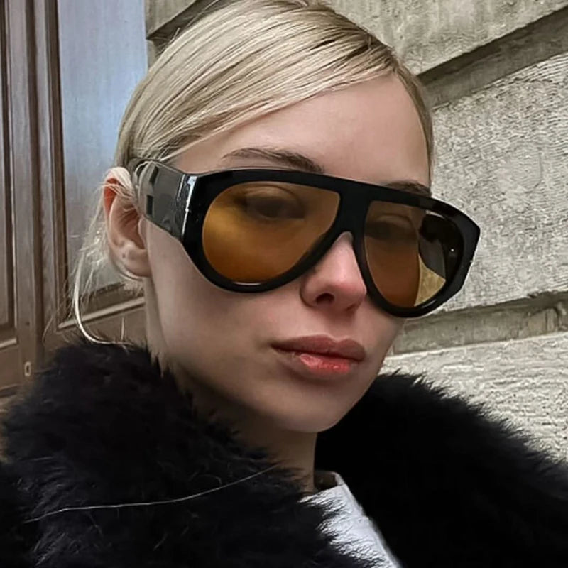 Delphine women’s shield sunglasses in Black frame with Amber lenses, worn by model against concrete wall — model facing slightly right, looking at camera, hands not visible — acetate frame, TAC lenses, 100% UV protection