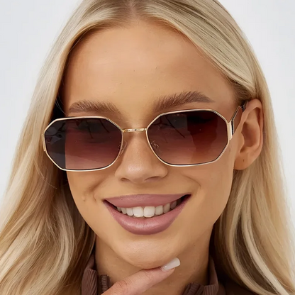 Model wearing Luna women’s retro geometric shades in Gold frame with Brown Gradient lenses, in front of white wall, facing forward and looking at the camera with a slight smile, left hand touching chin
