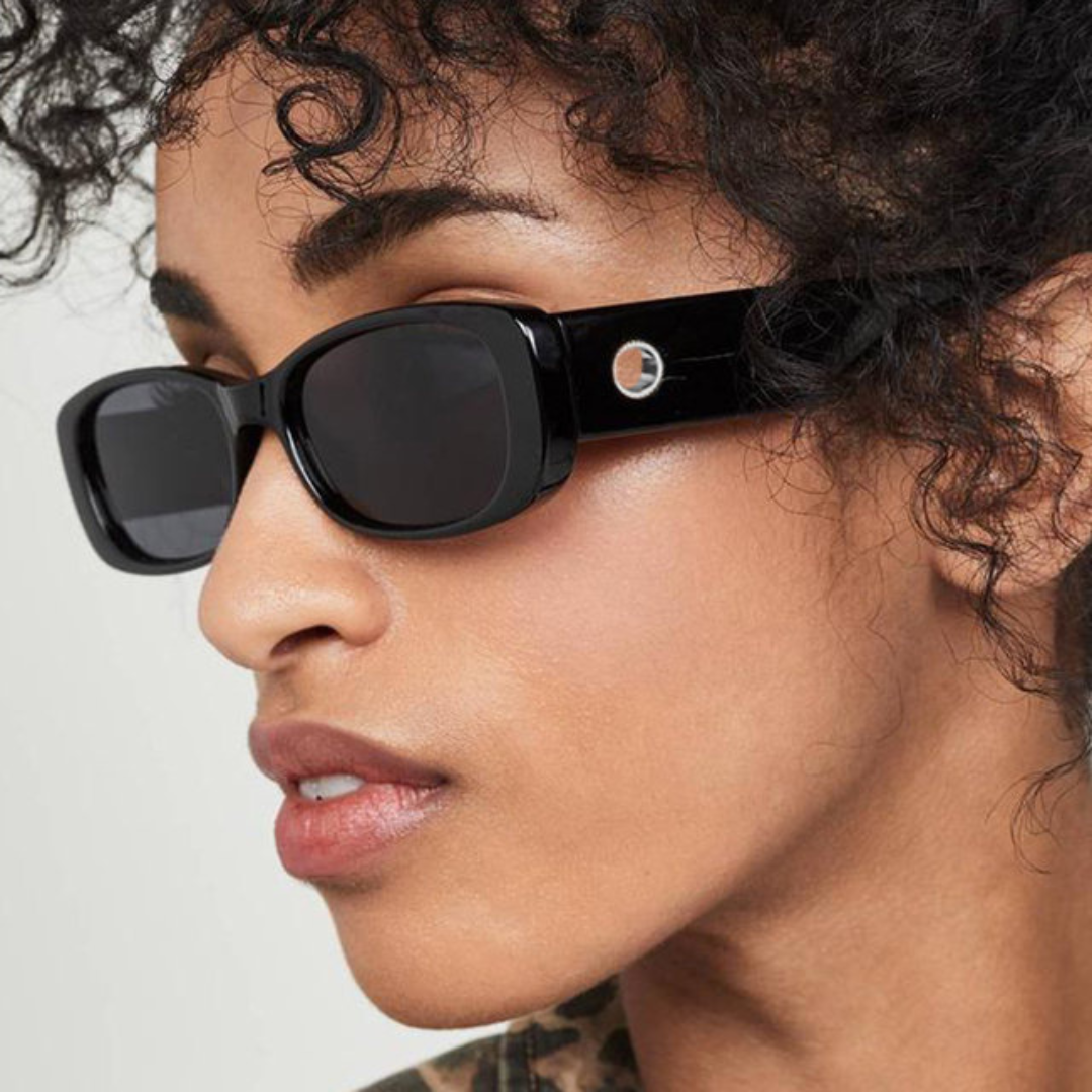 Milan women’s rectangular sunglasses in Black frame with Black lenses, worn by model against white wall — model facing left, looking left, hands not visible — premium acetate frame, Trivex lenses, 100% UV protection