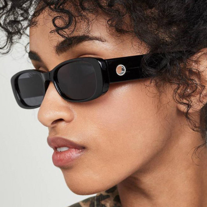 Milan women’s rectangular sunglasses in Black frame with Black lenses, worn by model against white wall — model facing left, looking left, hands not visible — premium acetate frame, Trivex lenses, 100% UV protection